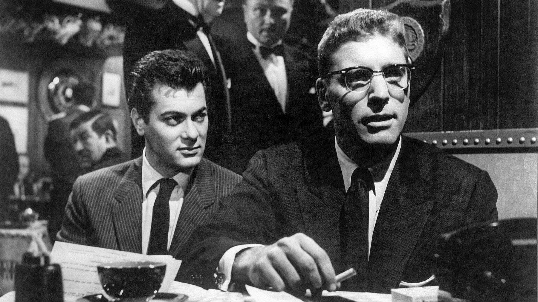 Tony Curtis and Burt Lancaster are sitting at a table having a coffee and cigarette and looking past us to someone out of the frame.
